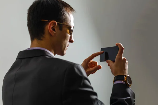 Νέοι χαρούμενα brunete δικηγόρος στέκεται στο καθαρό γαλάζιο φόντο και χαμογελώντας, διαβάζοντας καλά νέα στο διαδίκτυο στο κινητό του τηλέφωνο, φορώντας κοστούμι — Φωτογραφία Αρχείου