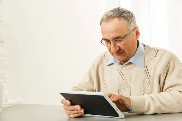 一位英俊而快乐的老人坐在客厅的桌子旁，他使用数字平板电脑。休闲活动、消磨时间、快乐退休和老年生活方式概念 — 图库照片