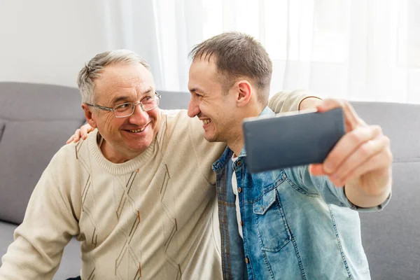 Lyckliga ögonblick. Glad ung man tar en selfie med sin optimistiska äldre far vinkade mot kameran och ler glatt — Stockfoto