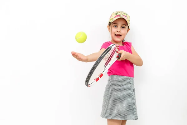 Ένα στούντιο σφηνάκι από ένα μικρό χαμογελαστή κορίτσι ντυμένο με ροζ T-shirt και λευκή φούστα-αθλητικά είδη για τένις σε λευκό φόντο. Στέκεται με μια ρακέτα του τένις και πετάει μια μπάλα του τένις — Φωτογραφία Αρχείου