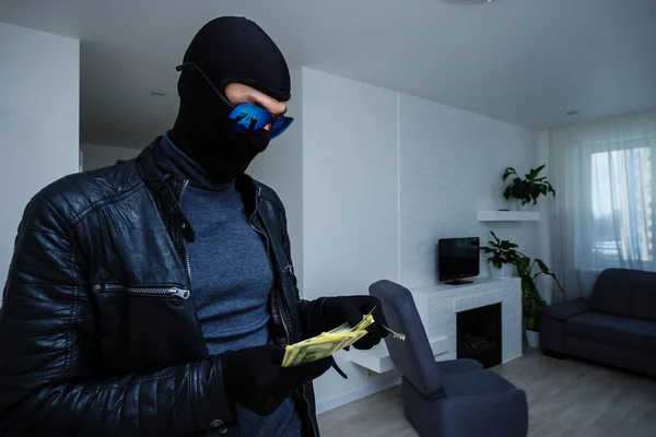 Robber man dressed in black hoodie stands with disguised face and holds a lot of money in his hands, stole a large amount, a thief man stole a TV — стоковое фото