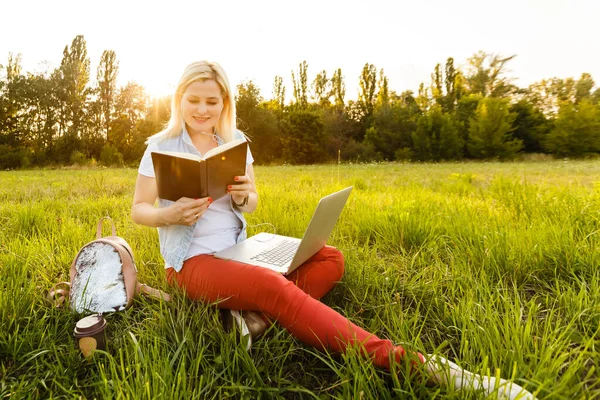 Jovem com laptop senta-se na grama no parque em um dia ensolarado. Loira rindo em um top de tanque branco e shorts. Treinamento online, trabalho remoto e comunicação em redes sociais — Fotografia de Stock