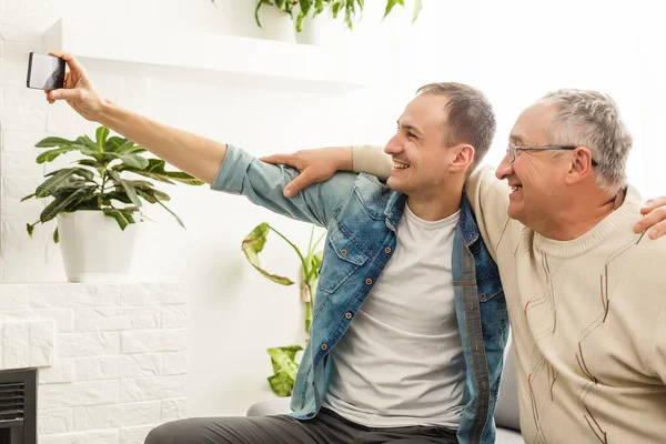 Um momento feliz. Jovem alegre tirando uma selfie com seu pai idoso otimista acenando para a câmera e sorrindo agradavelmente — Fotografia de Stock