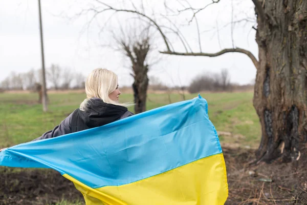 Kobieta trzyma żółto-niebieską flagę Ukrainy na tle spalonego drzewa — Zdjęcie stockowe