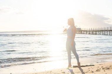 Sıska bir kadın, sakin deniz dalgalarının yanında yürüyor hafta sonunu doğada antrenman yapmak için geçiriyor eşofmanlı güzel bir kadın esnemek için uygun bir yer seçiyor.