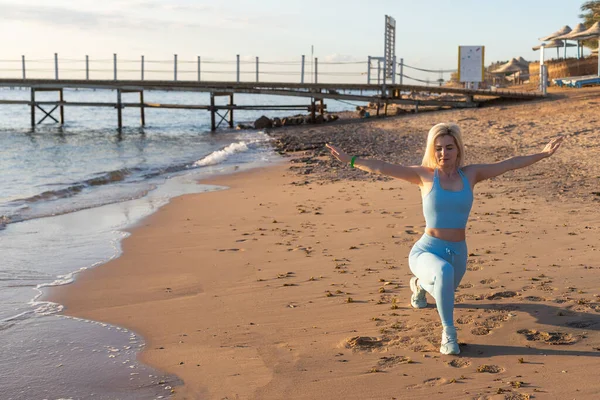 Attraente donna in forma che pratica affondi laterali, esercitandosi sulla spiaggia in una luminosa giornata di sole. Stile di vita sano, allenamento all'aperto — Foto Stock