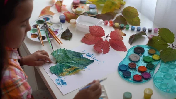 Close-up of little girl sitting at table drawing the leaves with brush and watercolours. little girl painting fallen leaves at home. autumn — Stock Photo, Image