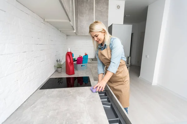 İnsanlar, ev işleri ve ev işleri konsepti. Evde mutfağı süngerle temizleyen bir şişe sprey ile sarışın orta yaşlı kadın.. — Stok fotoğraf
