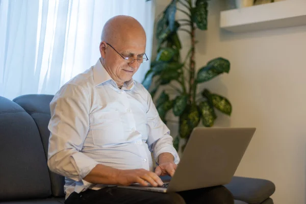 Beyaz gömlekli ve siyah pantolonlu yaşlı kel bir adam gözlük takıyor ve oturma odasında otururken dizüstü bilgisayarla çalışıyor.. — Stok fotoğraf
