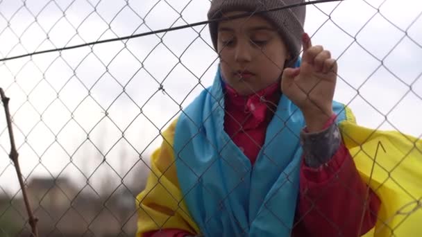 Metal bir çitin arkasında Ukrayna bayrağı taşıyan üzgün küçük bir kız. Mülteci ve zorunlu göçmen toplumsal sorunu — Stok video