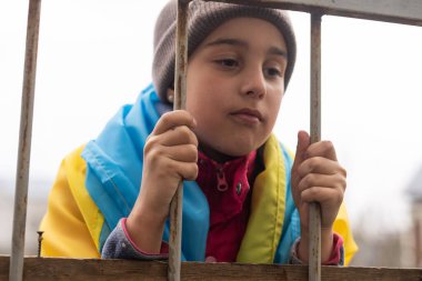 Metal bir çitin arkasında Ukrayna bayrağı taşıyan üzgün küçük bir kız. Mülteci ve zorunlu göçmen toplumsal sorunu