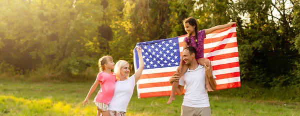 Happy family sitting together in their backyard holding the american flag behind them. Smiling couple with their kids celebrating american independence day holding american flag — Stock Photo, Image