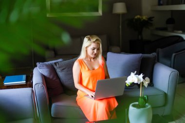 Modern bir otel lobisinde otururken bilgisayarla alışveriş yapan bir kadının yan görüntüsü. Elbise giyen sarışın bir kadın ekrana bakıyor.