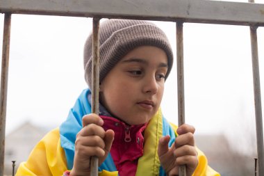 A little refugee girl with a sad look behind a metal fence. Social problem of refugees and internally displaced persons. Russias war against the Ukrainian people clipart