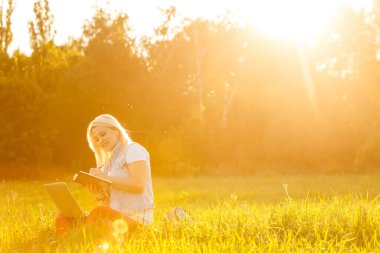 Dizüstü bilgisayarı olan genç bir kadın güneşli bir günde parkta çimlerin üzerinde oturur. Beyaz atletli ve şortlu, gülen bir sarışın. Çevrimiçi eğitim, uzaktan çalışma ve sosyal ağlarda iletişim