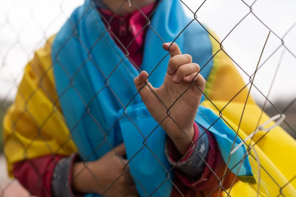 Smutná holčička s vlajkou Ukrajiny za kovovým plotem. Sociální problém uprchlíků a nucených migrantů — Stock fotografie