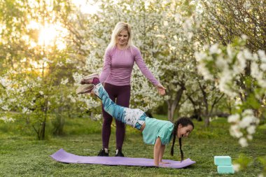 Genç sporcu anne, kızına yakın bahçede fizik egzersizi yapıyor. Sağlıklı bir yaşam tarzı. Yoga. Fitness. Aile dışarıda eğleniyor. Aile parkta birlikte vakit geçirir..
