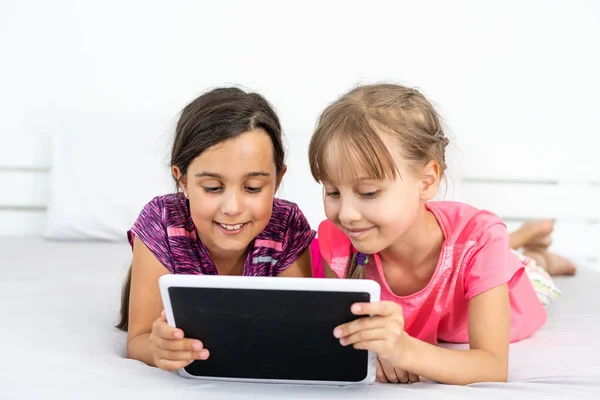 Dos niñas lindas están utilizando una tableta digital y sonriendo mientras están acostados en la cama en la habitación de los niños —  Fotos de Stock