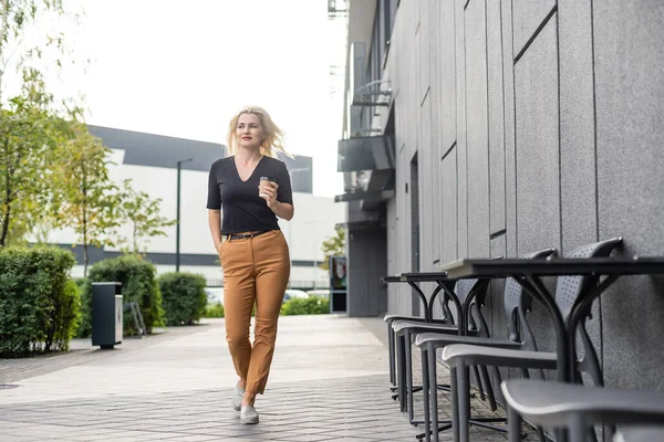 Hermosa mujer va a trabajar con café caminando cerca del edificio de oficinas. Retrato de una exitosa mujer de negocios sosteniendo una taza de bebida caliente en la mano en su camino al trabajo en City Street . — Foto de Stock