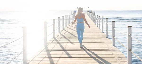 Vue latérale de la femme mince en vêtements de sport marchant près des vagues de mer calme passer le week-end matin pour l'entraînement à la nature, belle femelle en survêtement choisir l'endroit pour l'étirement — Photo