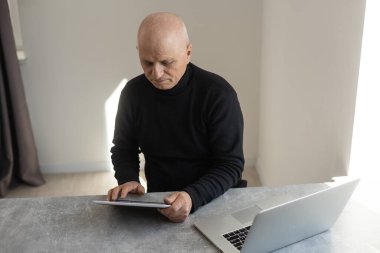 Kafası karışmış gri saçlı, kel, 60 yaşında, resmi giyinmiş, modern tableti karıştıran, şaşkına dönmüş bir şekilde masada oturuyor.