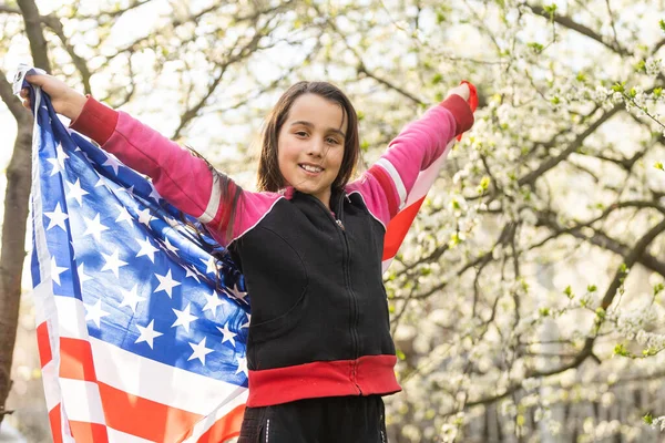 Счастливая очаровательная маленькая девочка улыбается и машет американским флагом снаружи. Улыбающийся ребенок празднует 4 июля - День независимости — стоковое фото