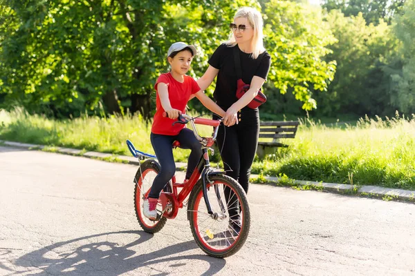 Όμορφη και ευτυχισμένη νεαρή μητέρα που μαθαίνει στην κόρη της να κάνει ποδήλατο. Και τα δύο χαμογελαστά, καλοκαιρινό πάρκο στο παρασκήνιο. — Φωτογραφία Αρχείου