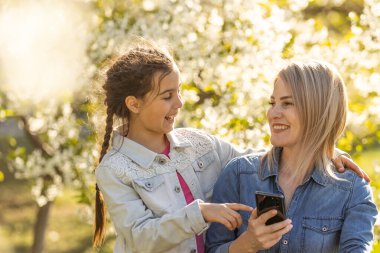 Mutlu kız ve anne dışarıda aletlerle rahatlıyor. Anne ve küçük kız akıllı telefonda ebeveyn kontrol uygulaması kullanıyor, online oyun oynuyor, dizüstü bilgisayarda video izliyor, internette geziniyor.