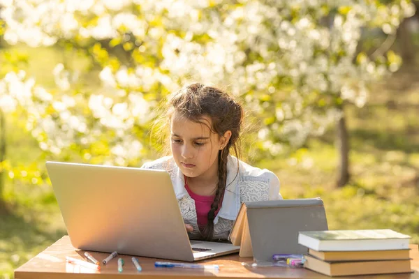 Školačka sedí venku, píše na klávesnici laptopu. Malá běloška sedící u stolu na verandě kavárny, studující s počítačem, dělá domácí úkoly. vzdálené učení, on-line vzdělávání — Stock fotografie