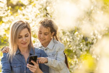 Mutlu kız ve anne dışarıda aletlerle rahatlıyor. Anne ve küçük kız akıllı telefonda ebeveyn kontrol uygulaması kullanıyor, online oyun oynuyor, dizüstü bilgisayarda video izliyor, internette geziniyor.
