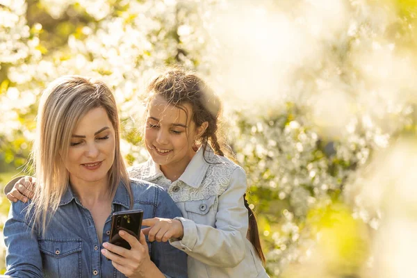 Happy girl and mom relaxing with gadgets outdoor. Mother and little daughter kid using parental control app on smartphone, playing online game, watching video on laptop, browsing internet