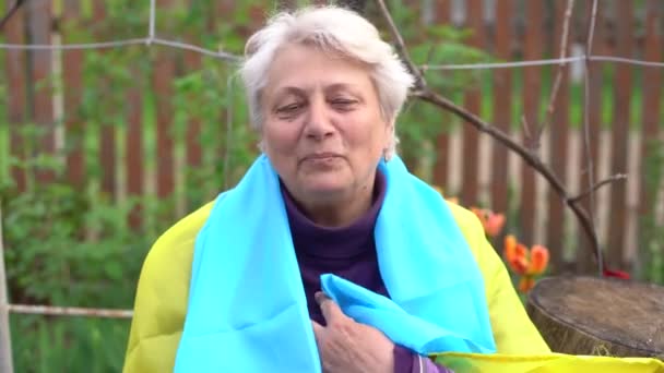 Senior white-haired woman wearing eyeglasses standing outdoors in the country waving the Ukrainian flag looking away feeling the freedom. No war, stop fights, we want peace. — Stockvideo