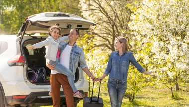 Happy family standing together near a car with open trunk enjoying view of rural landscape nature. Parents and their kid leaning on vehicle luggage compartment. Weekend travel and holidays concept.