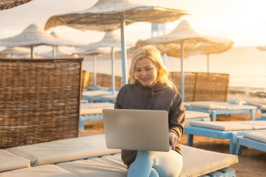 Yaz plajında gün batımında dizüstü bilgisayarlı bir kadın oturuyor.