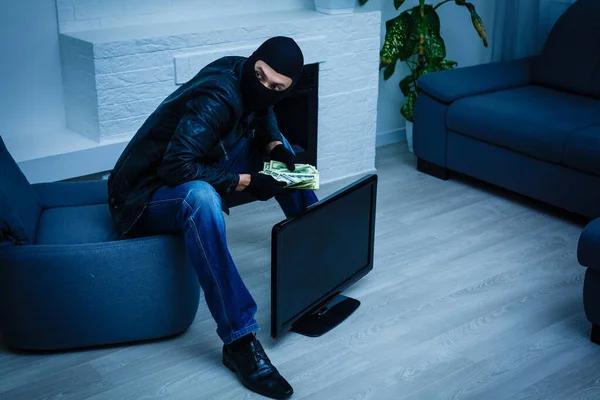 一个身穿黑色连帽衫的强盗站在那里 装模作样 手里拿着很多钱 偷了一大笔钱 一个小偷偷了一台电视机 — 图库照片