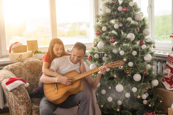 父と女の子はクリスマス休暇に一緒に楽しみを持っています 父は娘のためにギターを弾く 彼の小さい赤ん坊とのよい愛情のあるお父さんのレジャー 陽気な髭の男が歌を演奏 — ストック写真