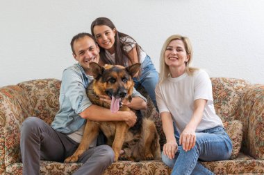 Güzel, genç ebeveynler, sevimli küçük kızları kameraya bakıyor ve gülümsüyor, evdeki kanepede sevimli köpekleriyle oturuyor..