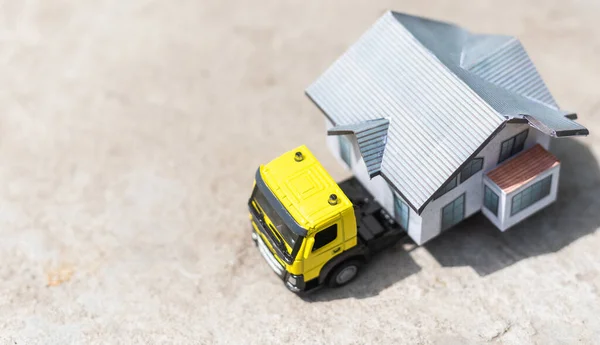 Close-up of a small toy truck on a tree stump carrying a miniature house model on top against a blurred forest background, front view. Countryside relocation service concept