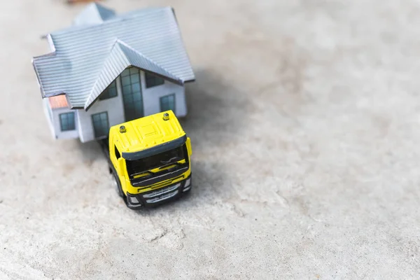 Close-up of a small toy truck on a tree stump carrying a miniature house model on top against a blurred forest background, front view. Countryside relocation service concept