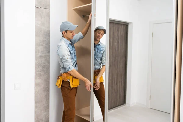 Employee furniture assembler in white t-shirt and jumpsuit checks wooden sliding door of modern wardrobe with mirror