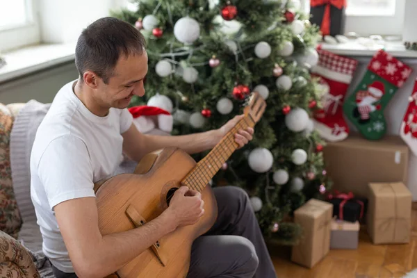 幸せな若い男がギターを弾いている 男は喜んで見ていると屈託のない クリスマスや新年を祝うだけでお祝いの帽子の男性 背景にガーランドとクリスマスツリー — ストック写真