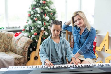 Mutlu Noeller ve mutlu tatiller. Neşeli anne ve tatlı kızı dekore edilmiş bir Noel ağacının arka planında beyaz bir piyanoda çalıyorlar. Yeni Yıl.