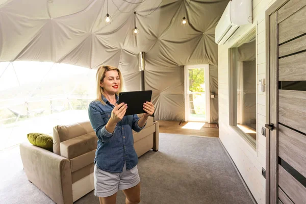 Young woman controlling home light with a digital tablet in the glamping dome tent. Concept of a smart home and light control with mobile devices.
