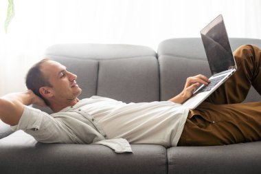 Genç ve çekici bir adam evdeki kanepede oturuyor, internette çalışıyor, internet kullanıyor, gülümsüyor, mutlu bir ruh hali içinde, serbest çalışıyor, serbest zaman geçiriyor, rahat, modern iş hayatı sürüyor.