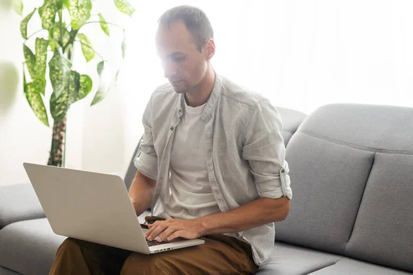 Young male tech user relaxing on sofa holding laptop computer mock up blank white screen. Man using modern notebook surfing internet, read news, distance online study work concept. Over shoulder view