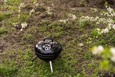 A metal barbecue grill stands in the backyard on the ground with grass by the bushes.