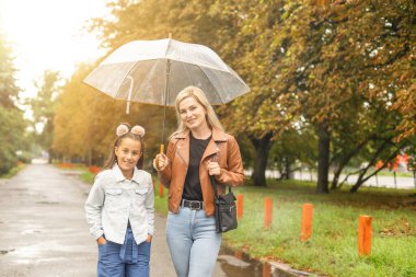 Mom holds a transparent umbrella from which fall autumn leaves. Daughter and mother laugh. Happy mom and little daughter having fun in autumn park
