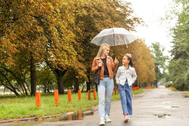 Mom holds a transparent umbrella from which fall autumn leaves. Daughter and mother laugh. Happy mom and little daughter having fun in autumn park