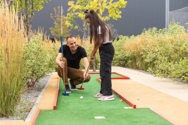 Sharing with golf experience. Cheerful young man teaching his daughter to play mini golf at the day time. Concept of friendly family.