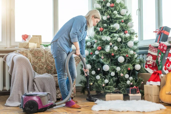 Young woman cleaning with vacuum cleaner, vacuuming under Christmas Tree needles with New Years ornaments on hardwood wooden floor